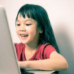 Enfant souriant face à son ordinateur sur lequel elle prend son cours en ligne
